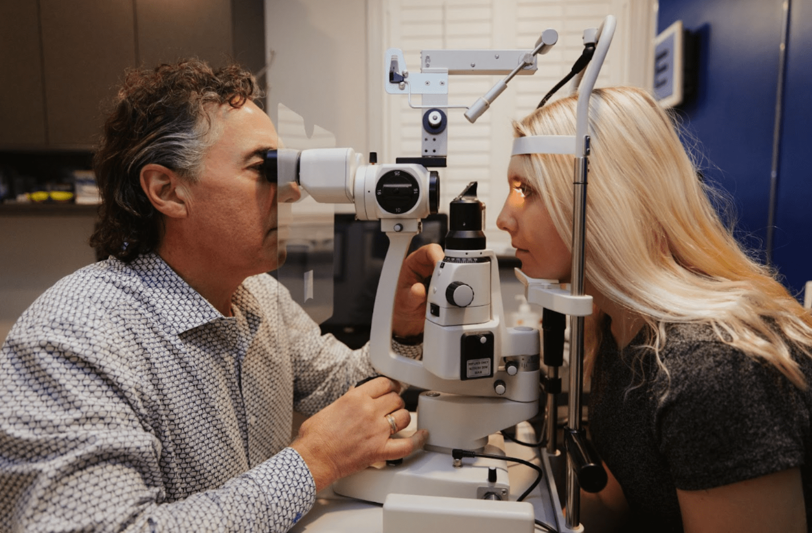 A female patient sitting behind a phoropter at the optometrist's office while Dr. Vagners examines her eyes