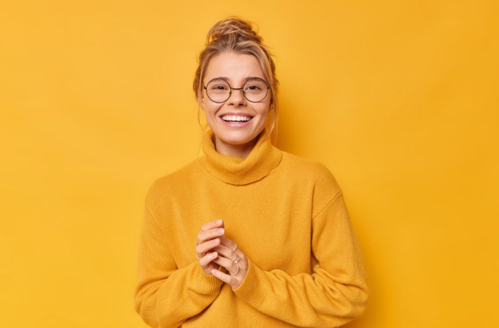 A young woman wearing a bright yellow sweater, also wearing glasses and smiling, leaning against a yellow background
