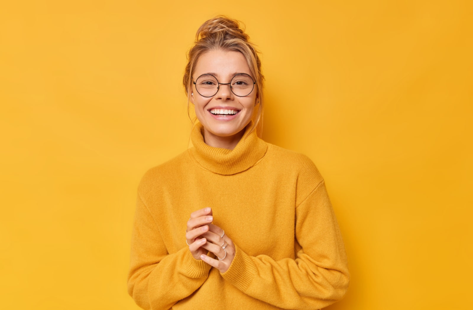 A young woman wearing a bright yellow sweater, also wearing glasses and smiling, leaning against a yellow background