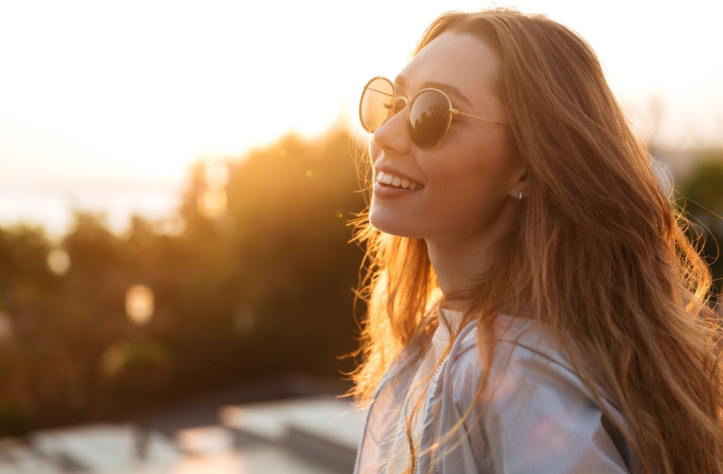 A young woman smiling towards the camera and wearing sunglasses to block the sun from her eyes