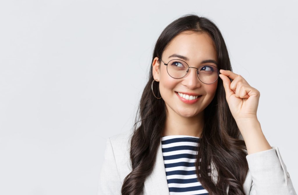 A woman smiling and looking up while she has one hand on her glasses