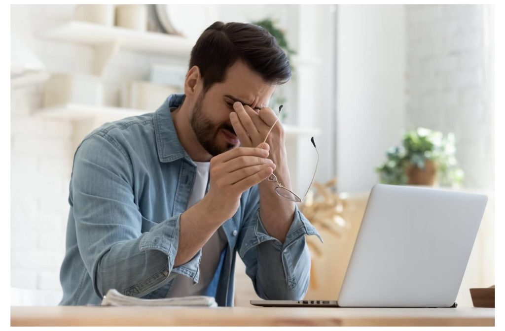 A young man taking off his glasses to rub his eyes while working due to dry eye syndrome.