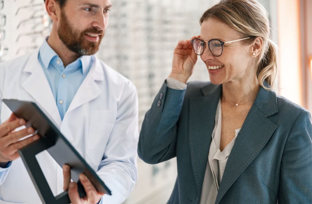 An optometrist holding a small mirror for a well-dressed woman as she smiles while trying on a new pair of glasses.