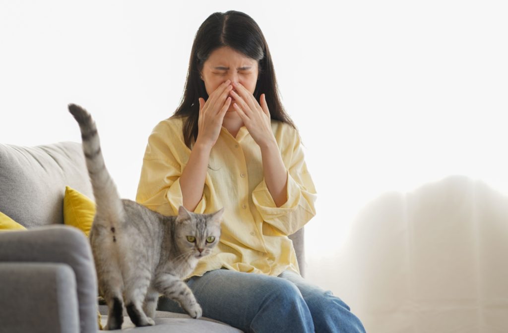 A young adult sitting on the couch beside their cat and rubbing their eyes in irritation.